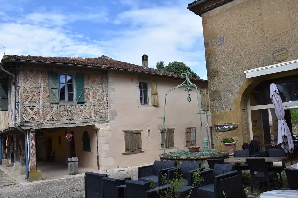 Place de village ancien avec une maison à colombages, des arcades en pierre et une terrasse sous un ciel bleu