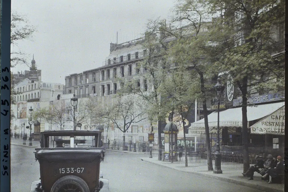 Photographie en couleurs anciennes d'un carrefour parisien bordé d'immeubles en pierre, avec une automobile des années 1920 au premier plan